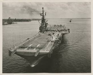 Black and white photograph of aircraft carrier USS Intrepid sailing with crew standing along po…