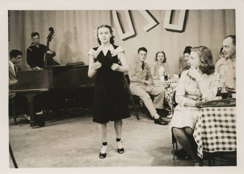 Black and white image of woman singing, a band is visible in the background, couples seated at …