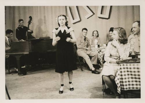Black and white image of woman singing, a band is visible in the background, couples seated at …