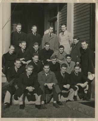 Black and white image of nineteen men wearing coats sitting and standing in rows next to a porc…