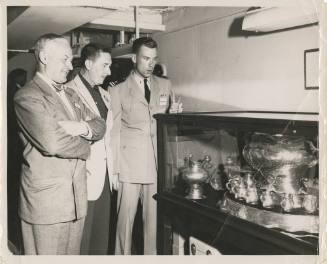 Black and white image of an officer showing two men a silver punch set with punch bowl and cups…