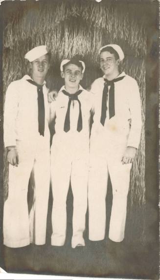 Black and white photograph of three sailors standing in front of a grass hut