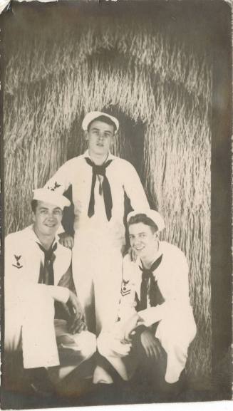 Black and white photograph of three sailors posing in front of a grass hut