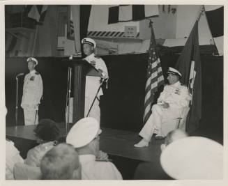 Black and white photograph of a change of command ceremony, with a naval officer speaking at a …