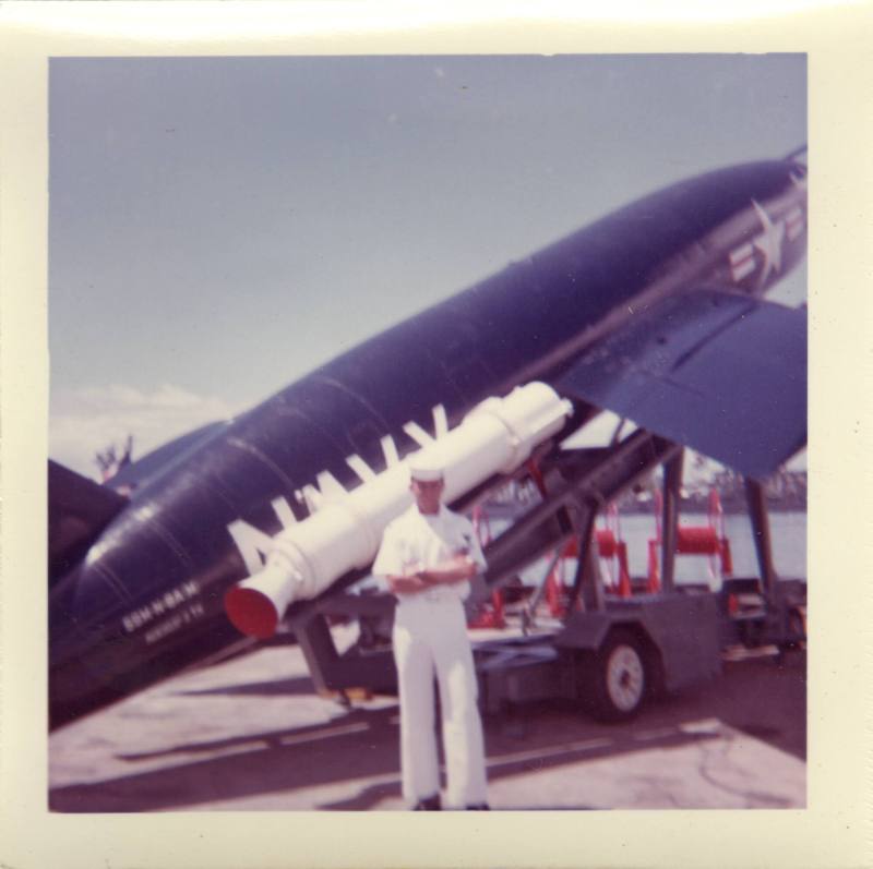 Color photograph of a sailor in a white uniform standing in front of a dark-blue Regulus I miss…
