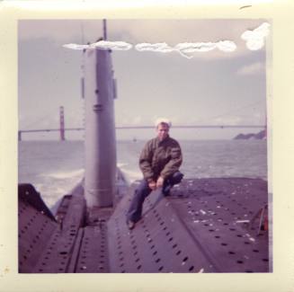 Color photograph of a sailor sitting on top of the submarine USS Growler, with the Golden Gate …