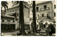 Black and white postcard of an outdoor cafe with palm trees and buildings in the background