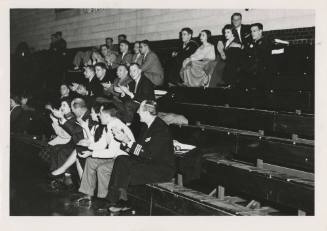 Black and white photograph of civilians and officers watching a game while sitting on bleachers…
