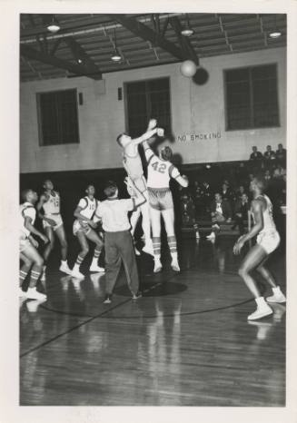 Black and white photograph of tip off during a basketball game with a referee standing in the m…