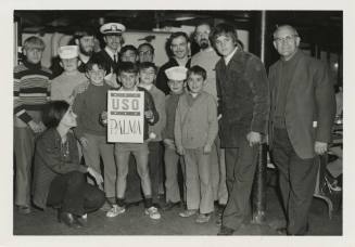 Black and white photograph of a group of civilians and children holding up a sign that says, “U…