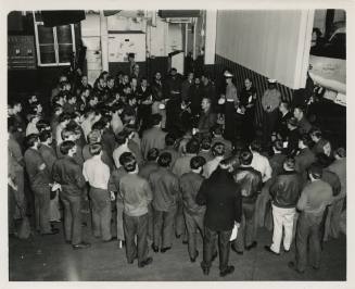 Black and white photograph of a group of men gathered on the hangar deck, listening to the comm…