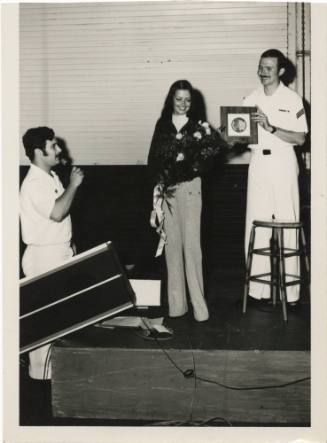 Black and white photo of two men and one woman standing on a stage, one sailor on the left spea…