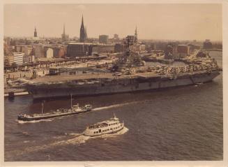 Color photograph of USS Intrepid docked in Hamburg, Germany, with buildings in the background