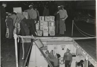 Black and white photograph of sailors transferring crates of supplies from the hangar deck thro…
