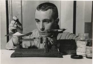 Black and white photograph of sailor measuring a white powdered chemical on a balance scale