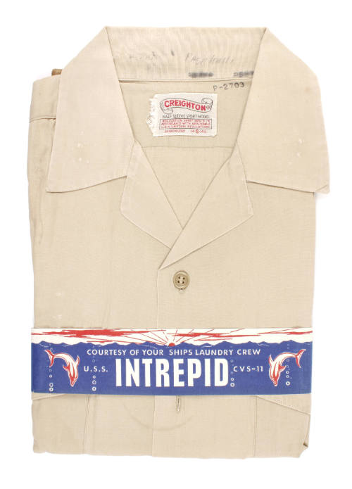 Folded khaki shirt with a blue paper band wrapped around that says "Intrepid, Courtesy of the s…