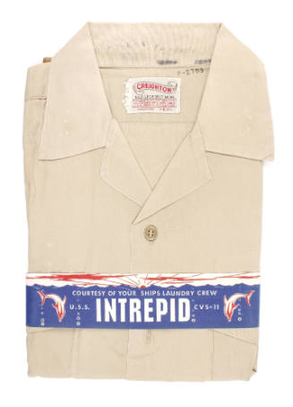 Folded khaki shirt with a blue paper band wrapped around that says "Intrepid, Courtesy of the s…