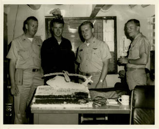 Black and white photograph of four U.S. Navy officers standing behind a desk with a helicopter …