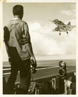 Black and white photograph of a man, with his back toward the viewer, watching a jet  land on t…