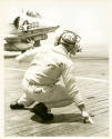 Black and white photograph of a catapult officer crouching on the flight deck, signaling a jet …