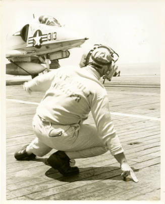 Black and white photograph of a catapult officer crouching on the flight deck, signaling a jet …
