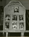 Black and white photograph of a wooden board with humorous portrait photographs of USS Intrepid…