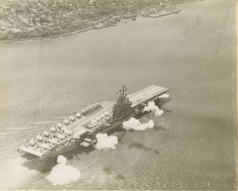 Black and white aerial photograph of USS Intrepid firing a salute with its anti-aircraft guns