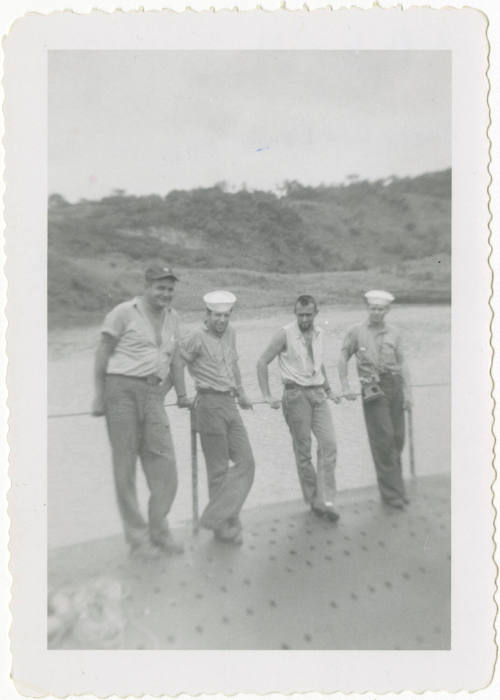 Printed black and white photograph of four sailors leaning on a railing on the top side of USS …