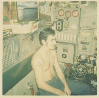 Printed color photograph of a shirtless man seated in USS Growler's engine room control booth