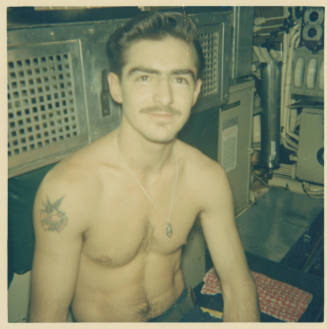 Printed color photograph of a shirtless man with dark hair and a mustache sitting in the engine…