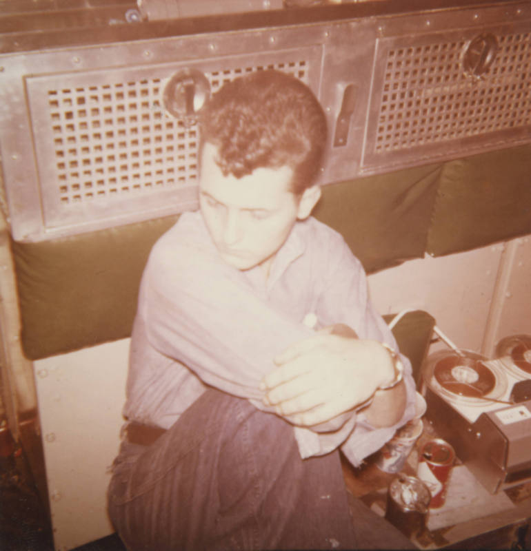 Printed color photograph of a man in a working dungaree uniform sitting in the engine room cont…