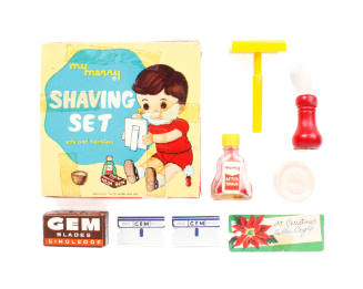 Square toy shaving set box surrounded by plastic razor and brush, disc of soap, aftershave, pap…