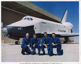 Color photograph of four men in blue flight suits posing in front of Enterprise