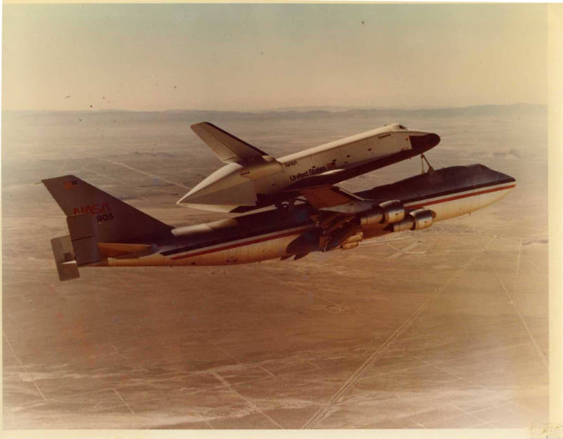 Color photograph of Enterprise atop airplane flying above flat land
