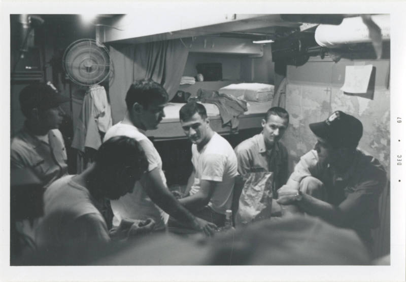 Black and white image of six men talking casually inside a stateroom 