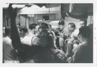 Black and white image of eleven men inside a stateroom, they are sitting on the bunks or standi…