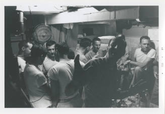 Black and white image of nine men inside a stateroom, they are standing or seated in circles ta…