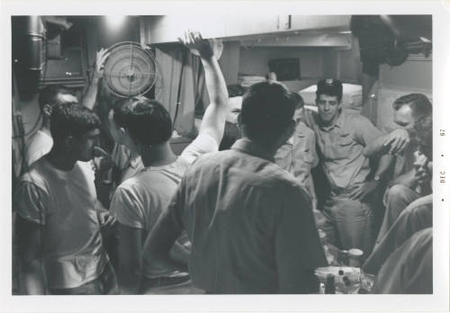 Black and white image of ten men inside a stateroom standing around talking, a table with bottl…