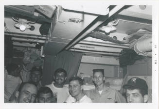 Black and white image of eight men standing inside a stateroom smiling for the camera