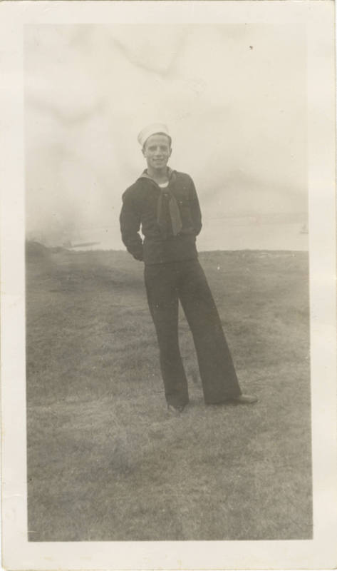 Black and white photograph of a sailor wearing a dress blue uniform and standing in a field