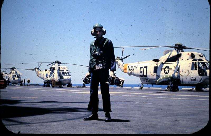 Color photograph of a sailor holding and wearing cameras on the flight deck with aircraft in th…