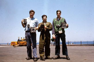 Color photograph of three photographer's mates holding cameras and posing on the flight deck