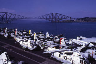 Color photograph of aircraft lined up on the flight deck with a bridge in the background