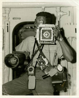 Black and white photograph of a photographer's mate holding many cameras in a ship's space