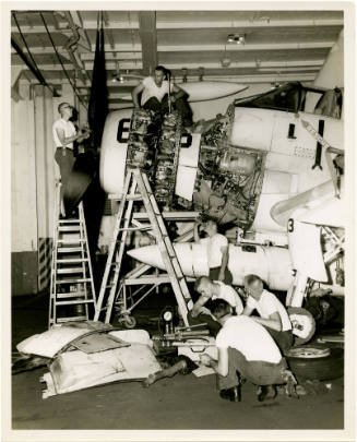 Black and white photograph of Intrepid crew members repairing an A-1 Skyraider in the hangar de…