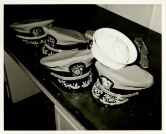 Black and white photograph of officer's caps on a desk with one sailor's "Dixie cup" hat on top…