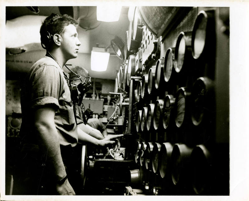 Black and white photograph of a crew member standing in front of gauges in an engine room