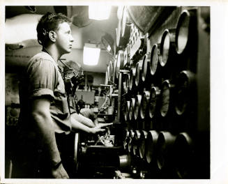 Black and white photograph of a crew member standing in front of gauges in an engine room