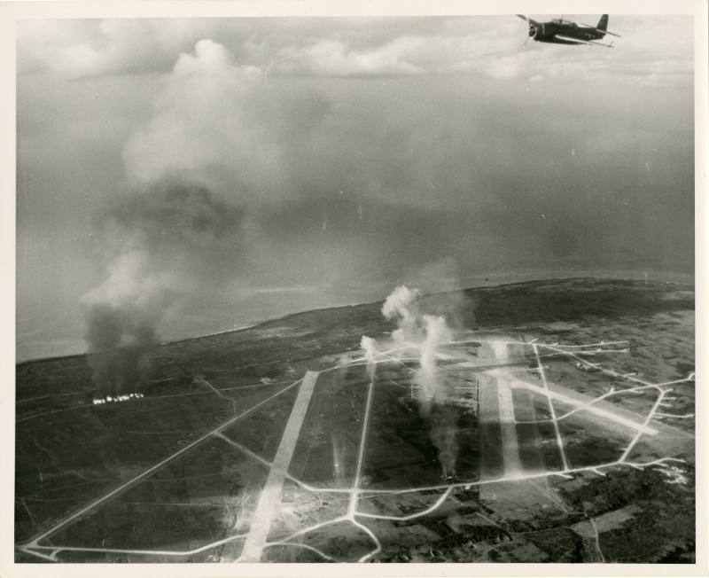 Black and white aerial photograph showing smoke rising from a bombed Japanese air base