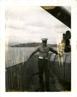 Printed black and white photograph of a Marine posing between an anti-aircraft gun and the rail…